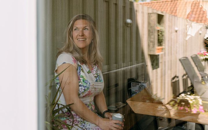 Blonde vrouw met een gebloemde jurk aan en een kop cappuccino in haar handen. Ze kijkt met een lach door een raam naar buiten. 