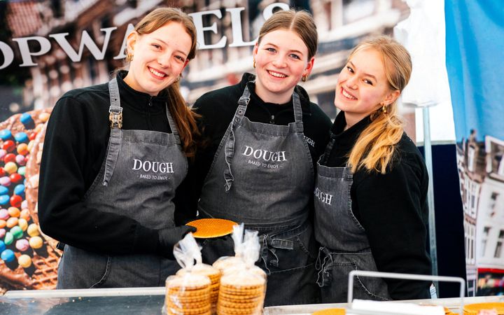 Drie jonge vrouwen in zwarte trui en grijze schort. Op de voorgrond pakjes met stroopkoeken.
