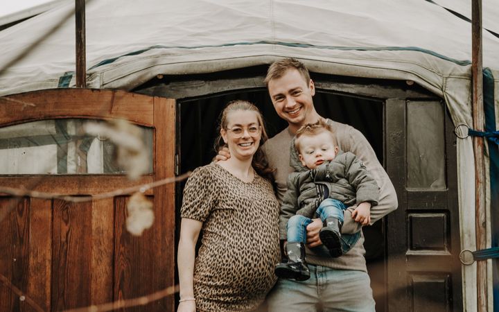 Jong paar, zij hoogzwanger en hij met een jong kind op de arm, voor de ingang van een nomadentent.