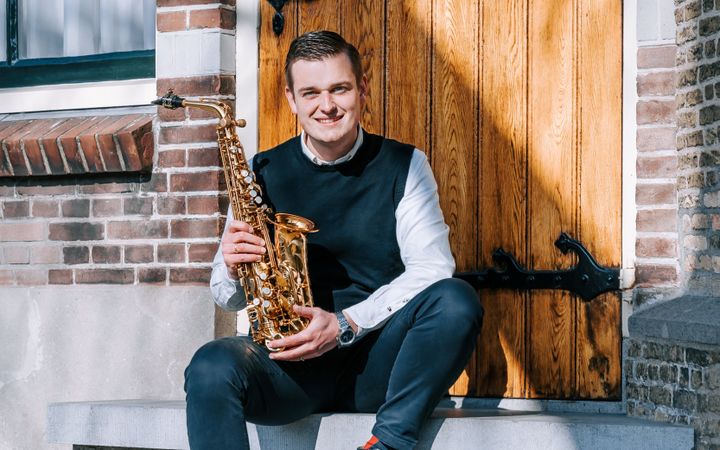 Jongeman in donkerblauwe spencer en licht overhemd, zittend op een stoepje, met een saxofoon in zijn handen.