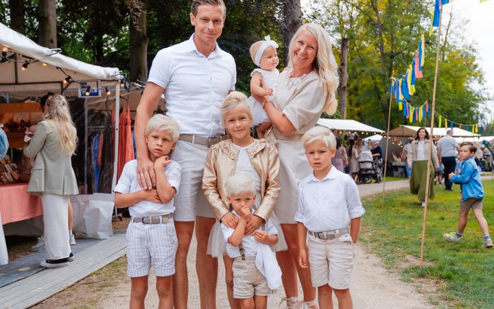 Gezin met vijf kinderen in lichte, zomerse kleding. Op de achtergrond stands van exposanten, in een parkachtige omgeving. Rechts kleurige vlaggetjes.
