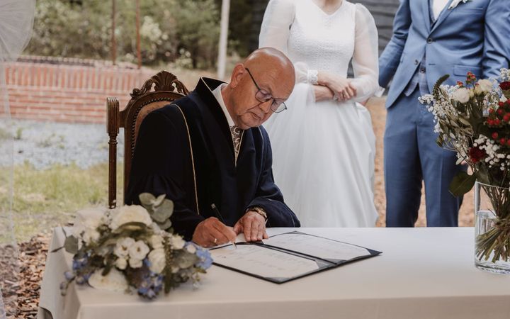 Gert van Tol zit in een toga achter een tafel met daarop een wit tafelkleed. Hij zet zijn handtekening in de akte voor de huwelijkssluiting van zijn kleinzoon en aangetrouwde kleindochter. Op de achtergrond is het bruidspaar deels zichtbaar.