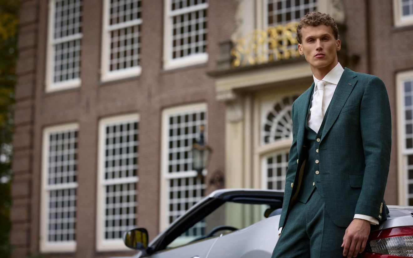 Bruidegom gekleed in groen trouwpak, staand naast een cabriolet voor een statig pand