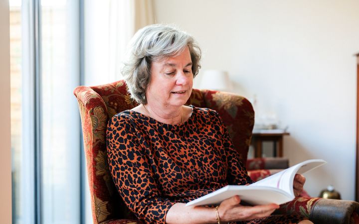 Vrouw met grijs haar en zwart met bruine jurk in een stoel, lezend in een boek.