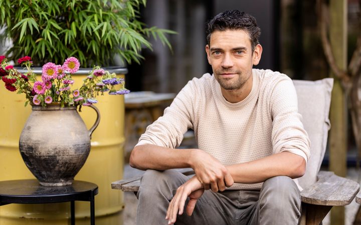 Jonge man met zwart haar en witte trui, zittend in een stoel naast een kan met bloemen.