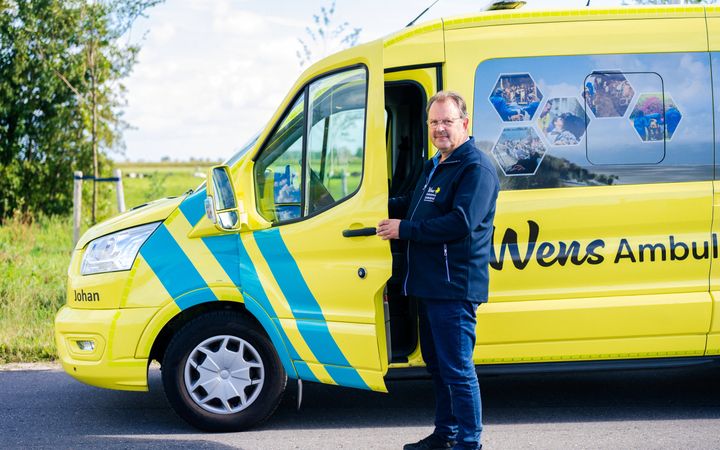 Een man in donkerblauwe kleding, bij een gele wensambulance.