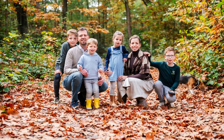 Een gezin in een herfstbos. Links een man in beige trui en jeans, op zijn hurken. Rechts een vrouw in beige rok, bruine coltrui en teddy vestje op haar hurken. Om hen heen vier jonge kinderen: drie jongens en een meisje.