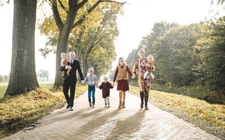 Man in zwart pak met jongetje op de arm en vrouw in kleurige jurk met meisje op de arm; ertussen drie oudere kinderen. Hand in hand op een landweg.
