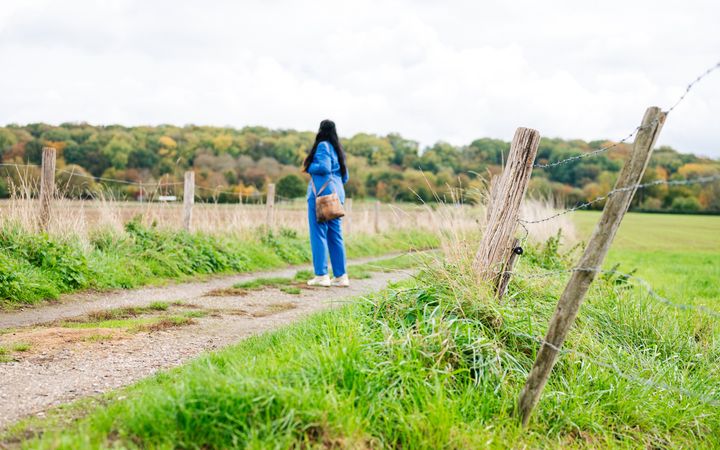 Vrouw in blauw broekpak, onherkenbaar gefotografeerd, in een landelijke omgeving met bos op de achtergrond.