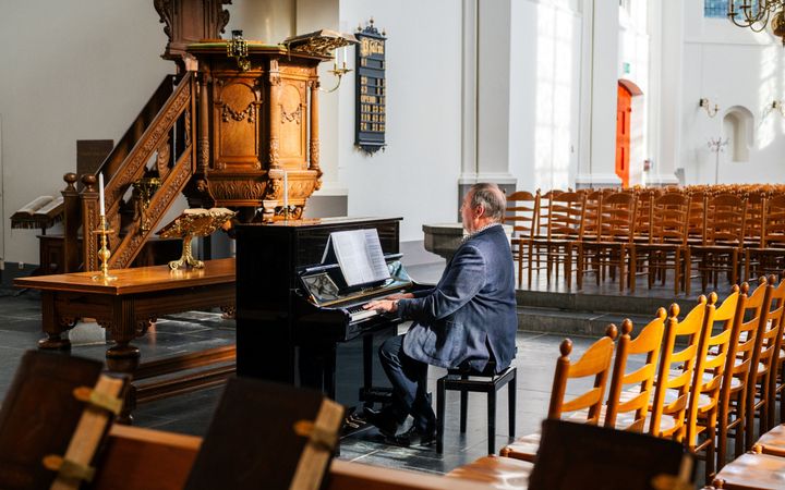 Een man in een blauw pak zit in een oude kerk bij een oude preekstoel piano te spelen.