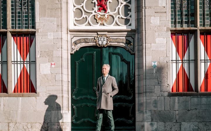 Man met grijs haar, groen kostuum en lichte overjas voor de deur van een monumentaal gebouw,