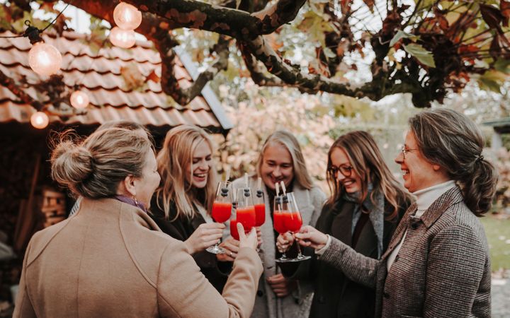 Vijf lachende vrouwen heffen champagneglazen met daarin rood sap, in een tuin met op de achtergrond een schuur.