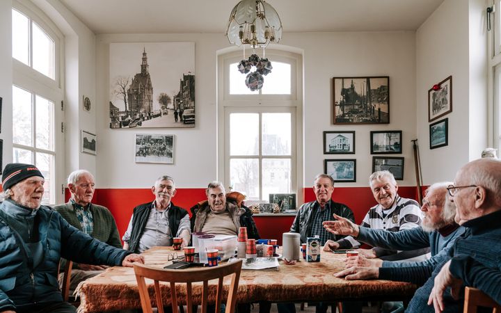 Oudere mannen, zittend rond een tafel in een kamer met een rood-witte muur en rondom vensters. Door het middelste raam is een toren te zien.
