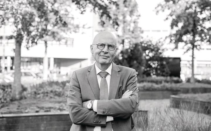 Portret van een man in pak, in een tuin bij een schoolgebouw.