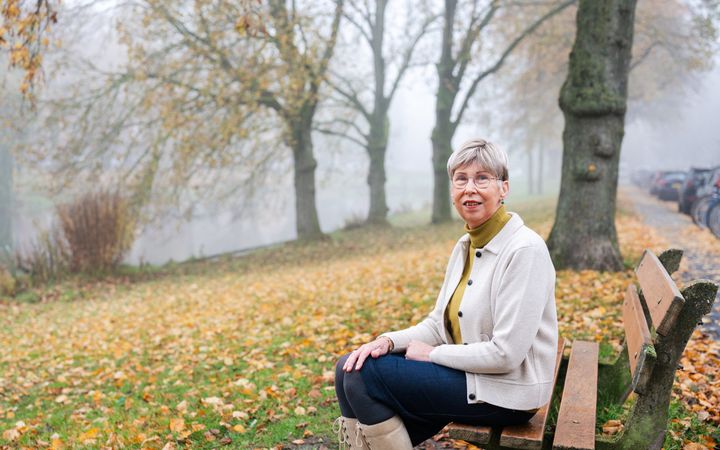 Vrouw met kort haar en een bril, in een groene coltrui, wit jasje en denim rok op een parkbankje. Op de achtergrond lichte mist, en bomen met wat geel blad. Voor haar een schuin aflopend grasveld vol met afgevallen blad. 