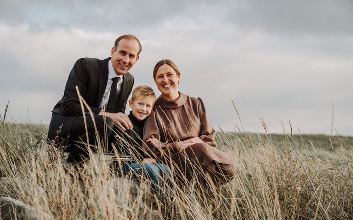 Man in donker pak met vrouw in een bruine jurk en tussen hen een blonde jongen in het gras. Op de achtergrond een grijze lucht.