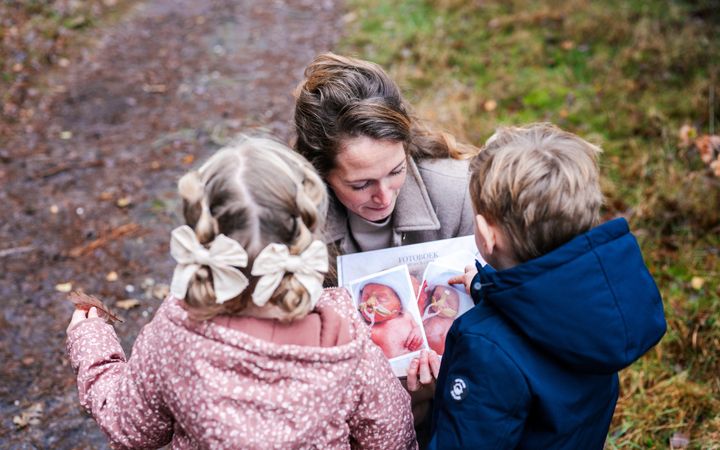 Jonge vrouw die op een bospad twee kleine kinderen een fotoboek laat zien met voorop de afbeeldingen van twee couveusekinderen.