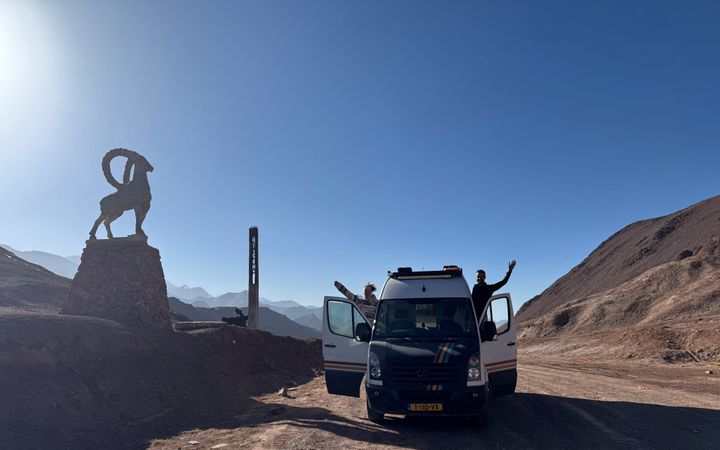 Camper op een onverharde weg tussen bergen. Een man en een vrouw zwaaien vanuit de geopende portieren.