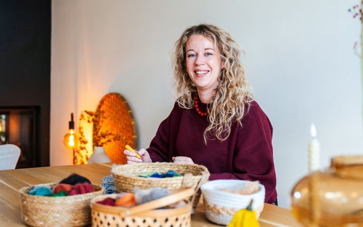 Vrouw met blonde krullen, een donkerrode trui en een rode kralenketting, zittend achter een houten tafel voor een witte muur. Op de voorgrond manden met gekleurde wol. 