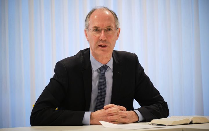 Man met dun grijs haar en blauw kostuum, zittend achter een tafel met gevouwen handen.