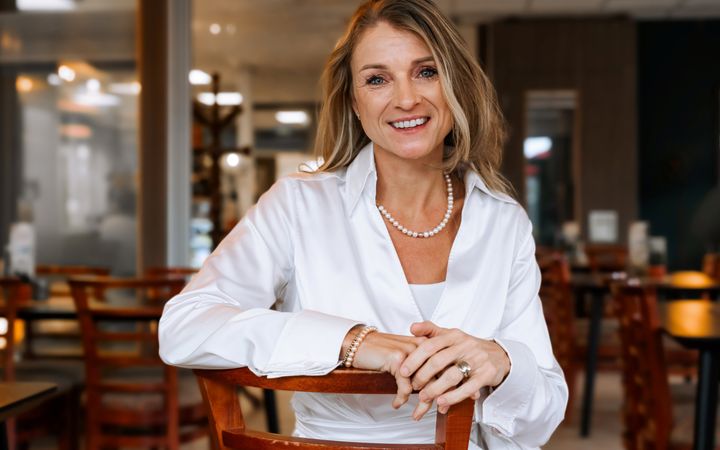 Burgemeester de Jonge, lang blond haar, witte blouse en parelketting, zit op een stoel in een Nijkerks ontmoetingscentrum.