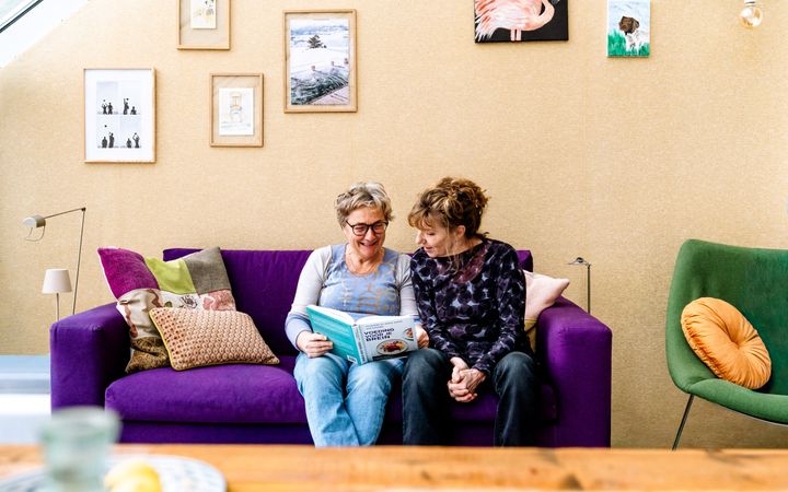 Twee vrouwen van middelbare leeftijd naast elkaar op een paarse bank, kijkend in een boek dat een van de vrouwen in de handen heeft.