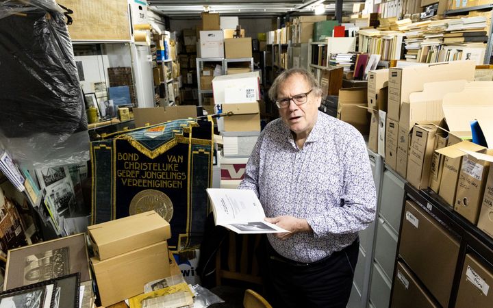 Man van middelbare leeftijd met een boek in de hand in een archiefruimte vol kasten, stellingen en dozen.