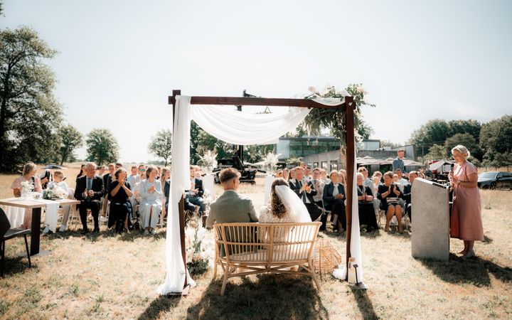 Vrouw in een roze jurk achter een katheder die in de openlucht een bruidspaar toespreekt.