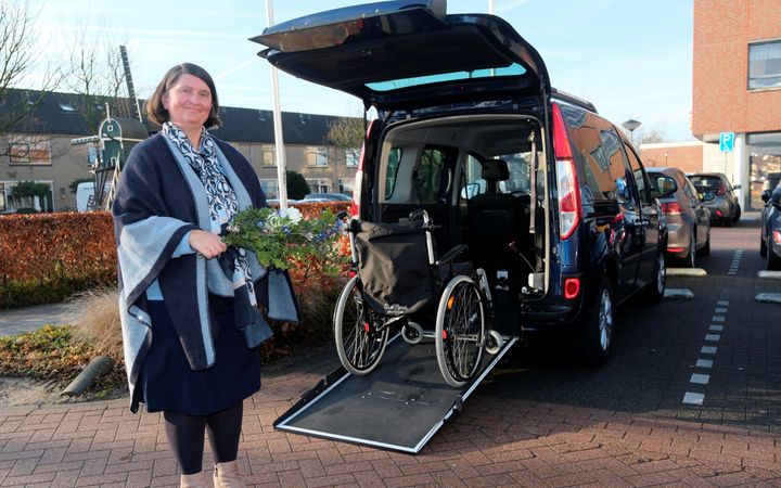 Vrouw met bruin boblijn-kapsel, donkerblauwe kleding en donkerblauwe omslagdoek met een boeket in de hand. Op de achtergrond een donkere bus met de klep open en een rolstoel op de rijplaat.