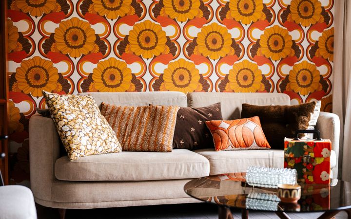 Beige bank vol kussens in oranje en donkerbruine tinten – tegen een wand met oranje-bruin-wit bloemenbehang.