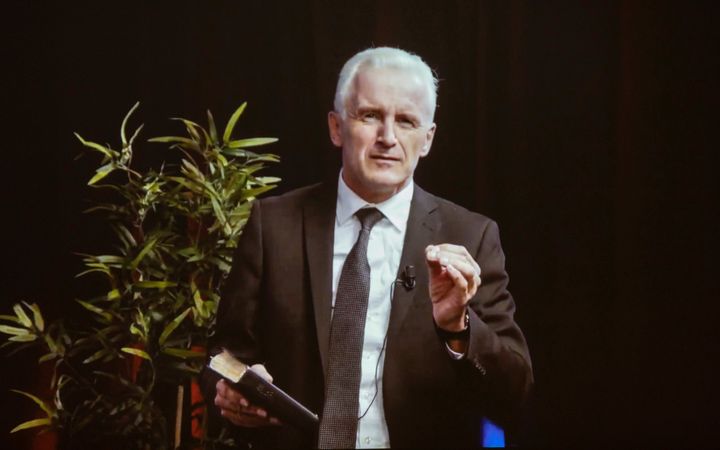 Man van middelbare leeftijd in donker pak, sprekend met opgeheven hand, in de andere hand een Bijbel.