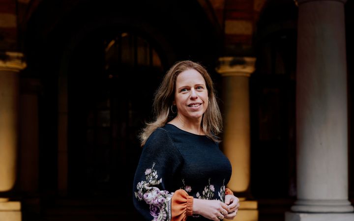 Burgemeesster Carola Schouten staat naast een pilaar in het gemeentehuis van Rotterdam. Ze heeft lang, blond haar en een zwarte gebloemde jurk aan.