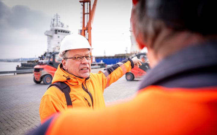 Man in oranje jas en witte helm op het hoofd in gesprek op een kade met op de achtergrond een zeeschip.