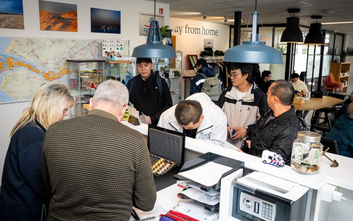Zaal met buitenlandse mensen, achter de balie een Nederlandse man en vrouw.
