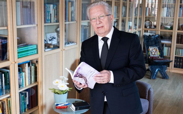 Man met bril, grijs haar en donker pak voor een boekenkast met een boekje in de hand.