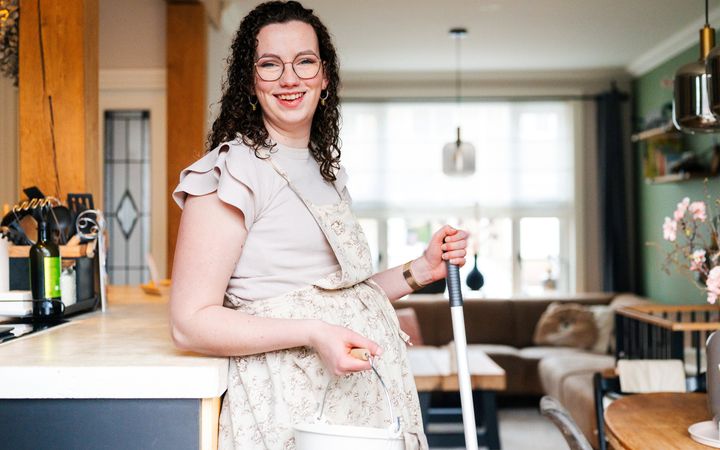 Aline van den Boogaard (donkere krullen, bril, beige kleding) heeft een dweilstok in haar handen.