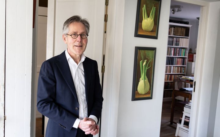 Man van middelbare leeftijd in donkerblauw kostuum in de deuropening van een kamer met boekenkast en schilderijen aan de wand.