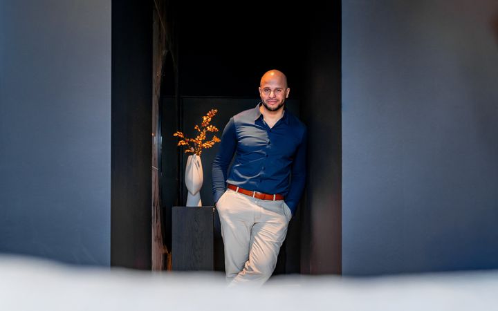 Man in blauw overhemd en lichte broek, leunt tegen een blauwe muur. Links van hem een vaas met een tak.