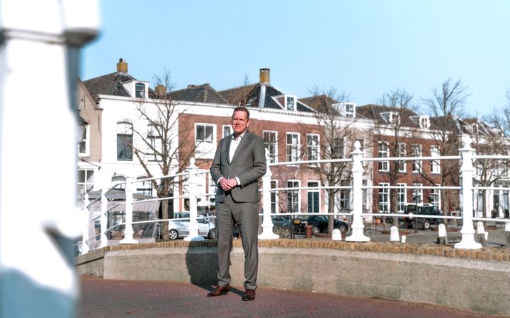 Man in grijs pak, staand op een brug, met op de achtergrond historische gevels.