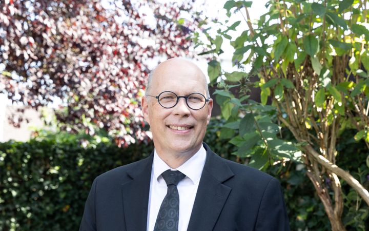 Man met kaal hoofd, ronde bril en donker pak in een tuin met heesters.