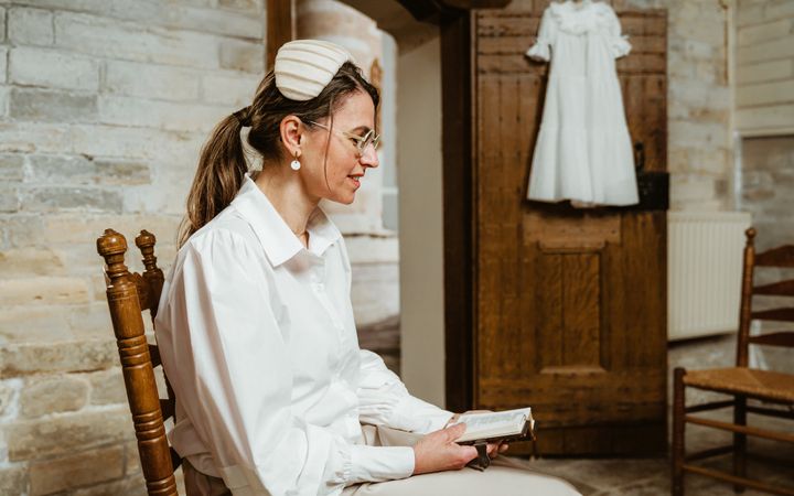 In het wit geklede vrouw met wit hoedje, zittend in een kerk waar ze uit een Bijbeltje leest. Aan een kast naast haar hangt een doopjurk.