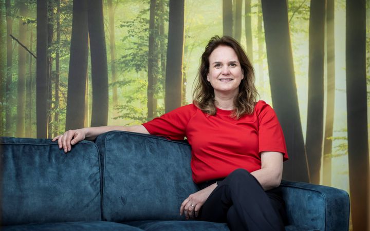 Donkerblonde vrouw, met een helderrood shirt aan op een blauwe bank. Op de achtergrond een wandvullende foto van een bos. 