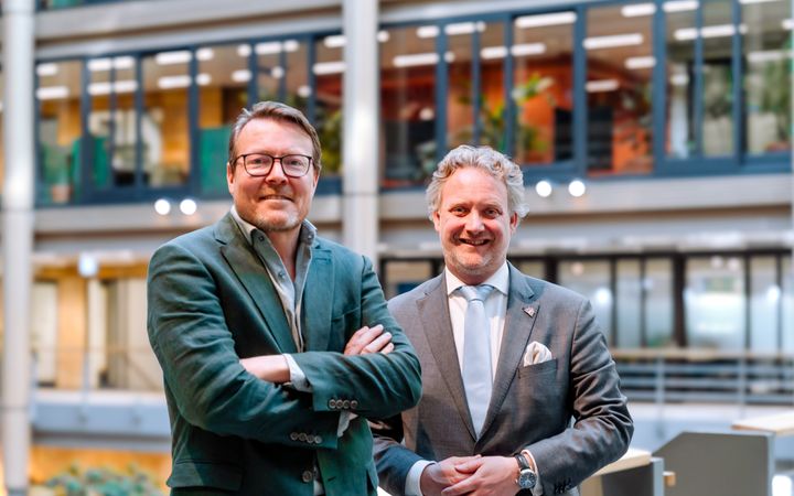 Man in groen pak en man in grijs pak, staand in een groot kantoorgebouw.