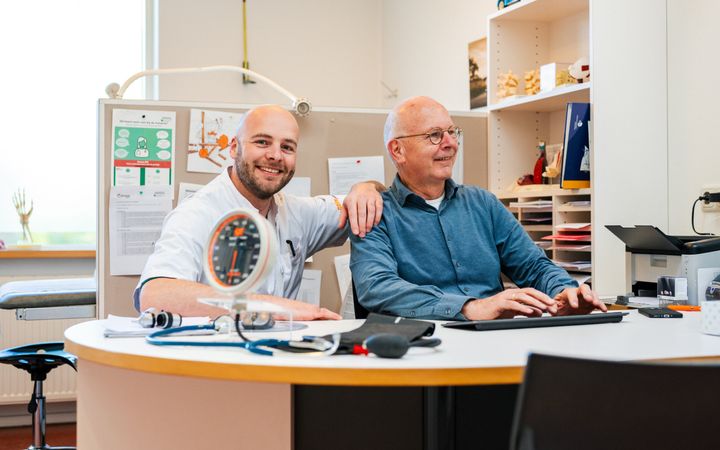 Jonge man in witte doktersjas naast oudere man met blauw overhemd aan een bureau.