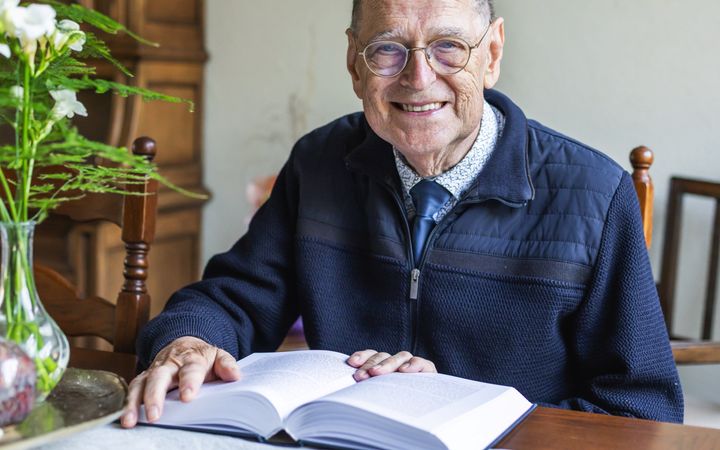 Oude man met een blauwe trui aan een tafel met een boek voor zich.
