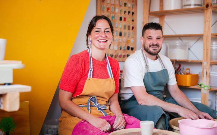 Links een lachende vrouw met donker haar en een oranje schort om, rechts een man met donker haar en een blauw short voor. Beiden zitten voor een pottenbakkersschijf. 