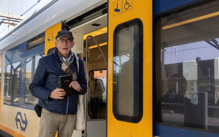 Man met bril op, beige broek en donkerblauwe jas en pet, met een zwarte Bijbel in zijn handen; staande voor de geopende deuren van een NS-trein. 