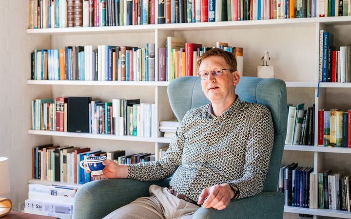 Man van middelbare leeftijd met rood haar voor een boekenkast met een koffiebeker in de hand.