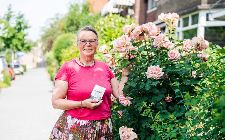 Vrouw van middelbare leeftijd met roze shirt en gebloemde jurk voor een rozenstruik, in haar rechterhand het zwartwitportret van een oudere man.