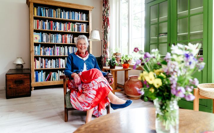 Vrouw met grijs haar, rode rok en blauw vestje, zittend op een stoel in een woonkamer.